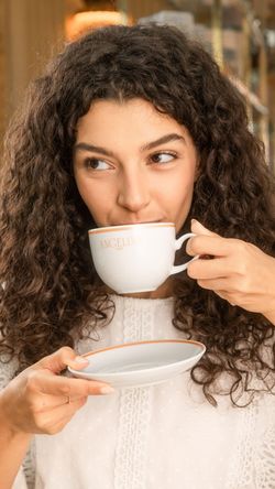 Woman drinking hot chocolate at Angelina Paris - Rivoli tea room, holding a branded cup and saucer.