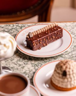 Chocolate-covered pastry topped with cream on an Angelina Paris plate, accompanied by a mont-blanc dessert and hot chocolate.
