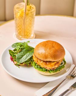 Chicken burger with lettuce and mixed greens on a white plate, served with a glass of iced lemon drink.