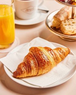Golden croissant on a white plate with a napkin, accompanied by a glass of orange juice and a pastry stand with bread slices.
