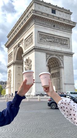 Deux gobelets de chocolat chaud Angelina Paris - Rivoli tenus devant l'Arc de Triomphe à Paris.