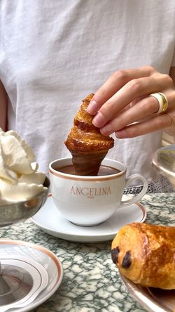 Hand dipping a croissant into a cup of hot chocolate at Angelina Paris tea room, with pastries on the side.