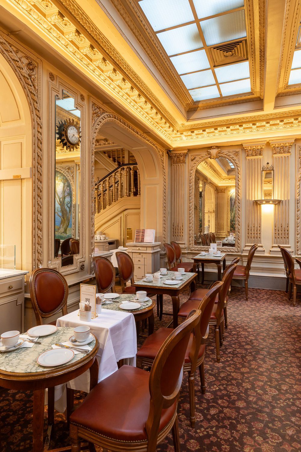 Salon de thé ancien avec tables en marbre, chaises en cuir, décor doré et éclairage naturel par puits de lumière.