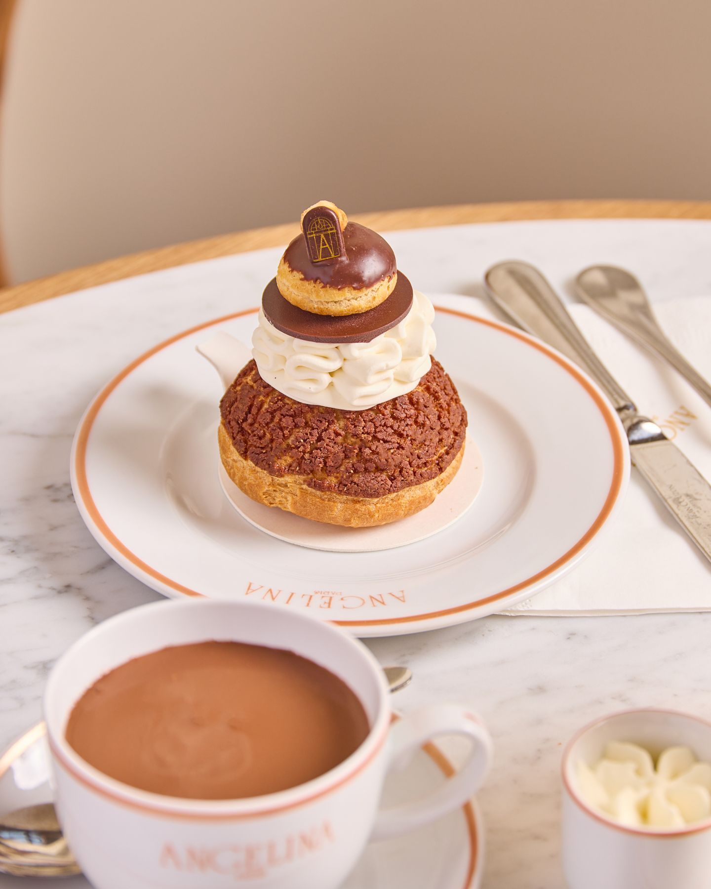 Pastry with chocolate topping and whipped cream on a plate, served with a cup of hot chocolate and butter on a marble table.