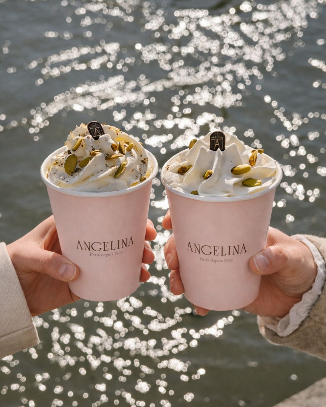 Two cups of hot chocolate with whipped cream and pistachios held in front of sparkling water at Angelina Paris tea room.