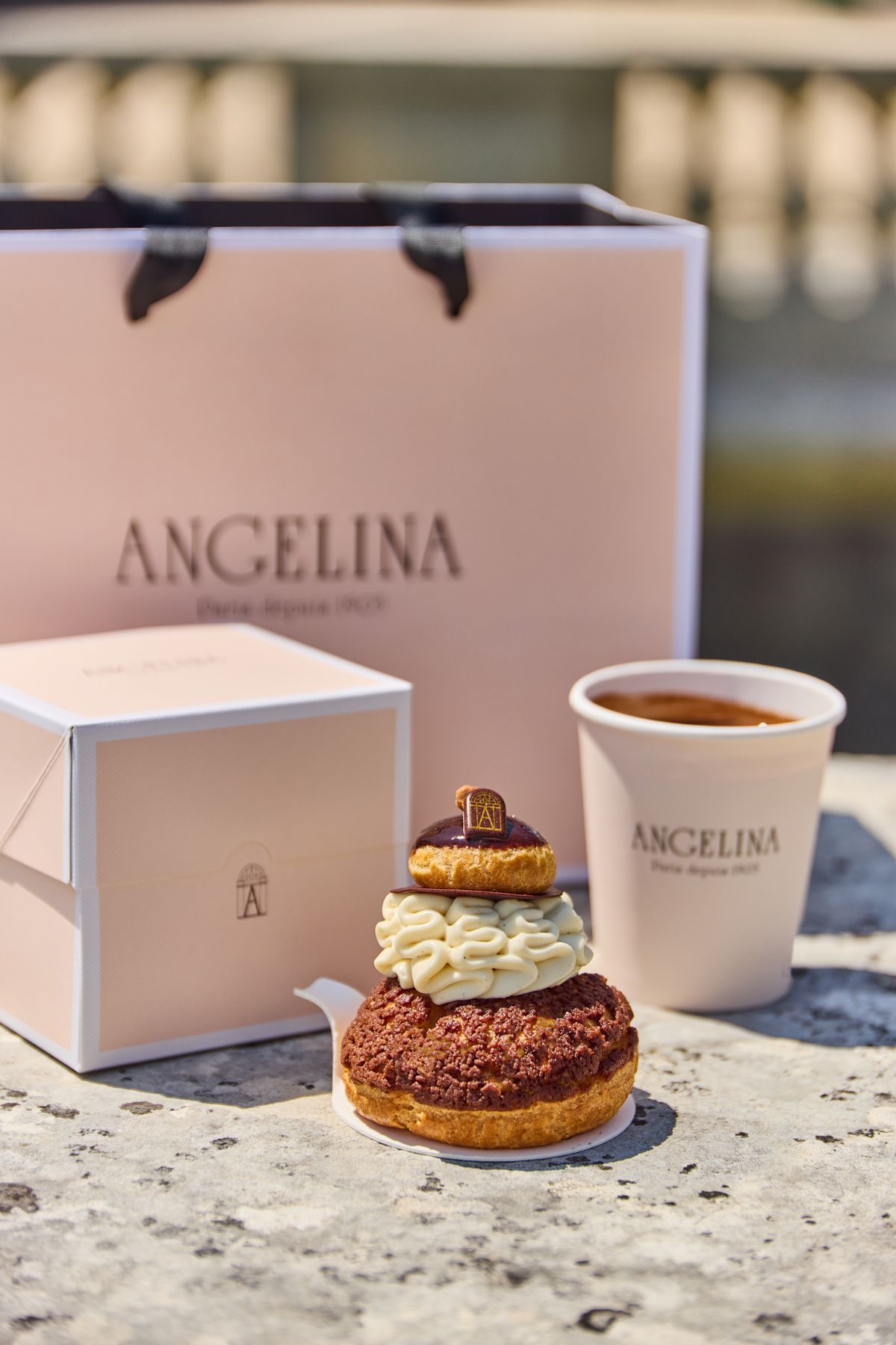 Pastry and hot chocolate from Angelina Paris - Domaine de Trianon displayed with branded packaging and cup outdoors.
