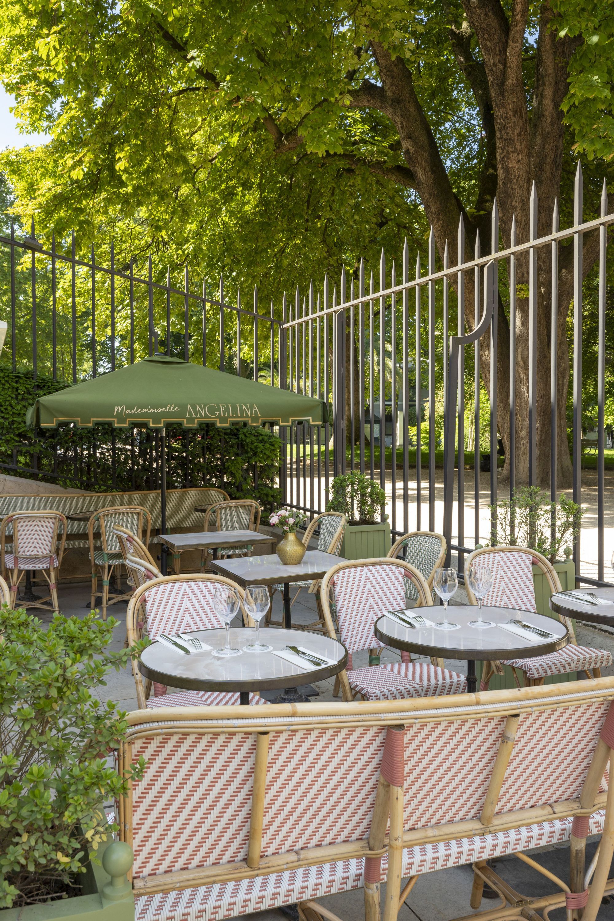 Outdoor seating area at Mademoiselle Angelina tea room with tables, chairs, and green parasols surrounded by trees and fence.
