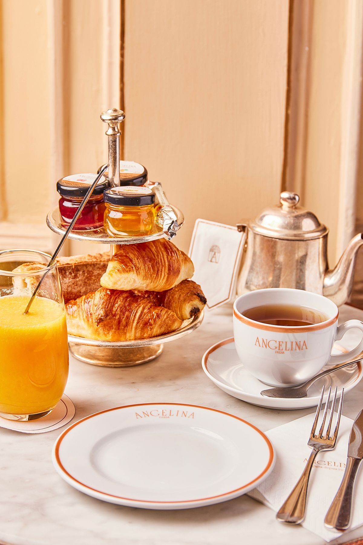 Breakfast setup at Angelina Paris - Rivoli with croissants, jam jars, orange juice, and a cup of tea on a marble table.