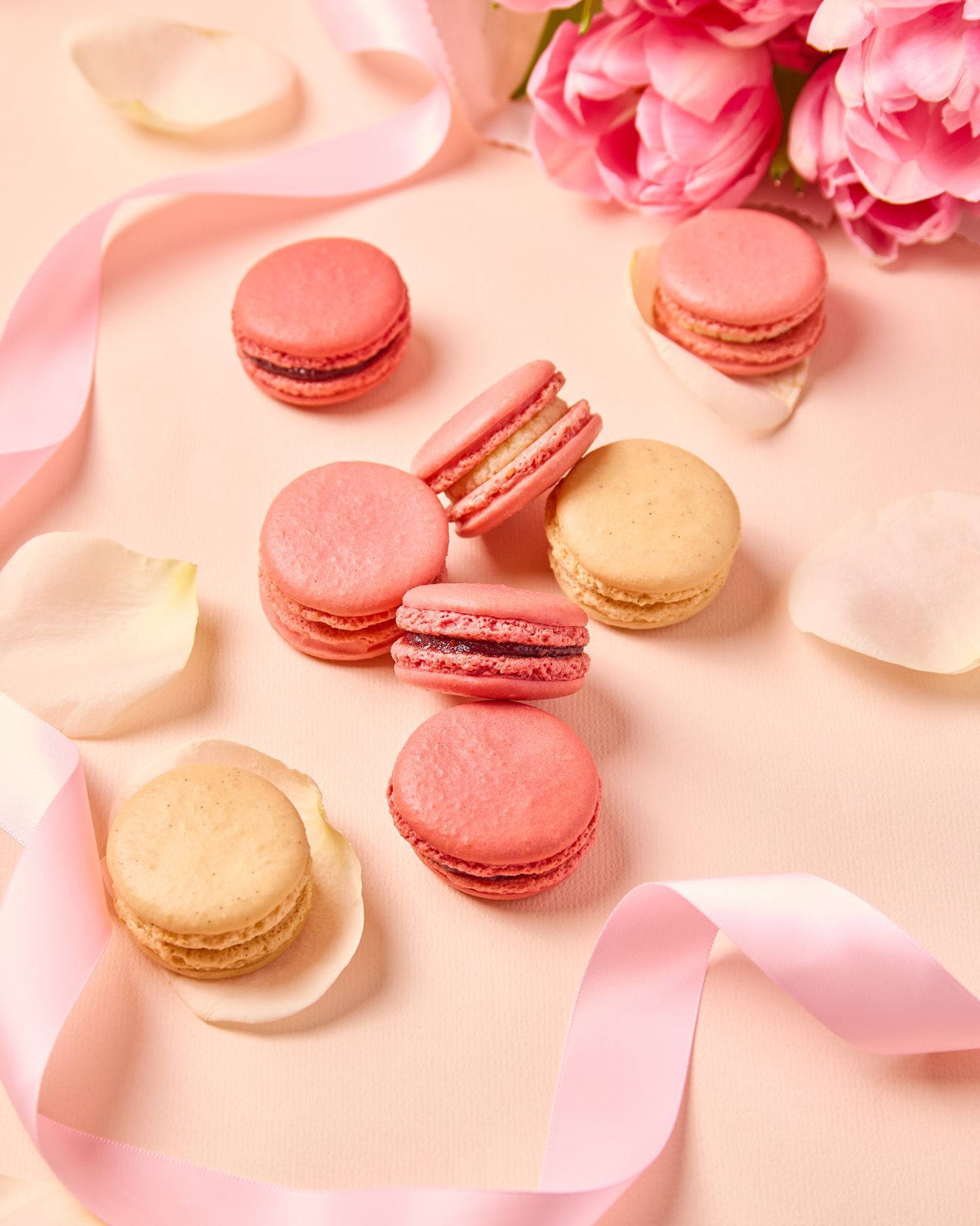 Pink and beige macarons arranged with rose petals and pink tulips on a light pink surface with a satin ribbon.