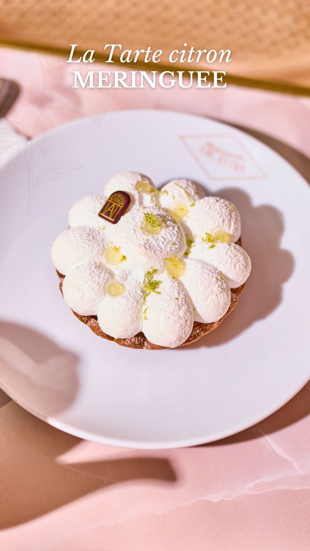 Lemon meringue tart served on a white plate with Mademoiselle Angelina branding on a pink surface.