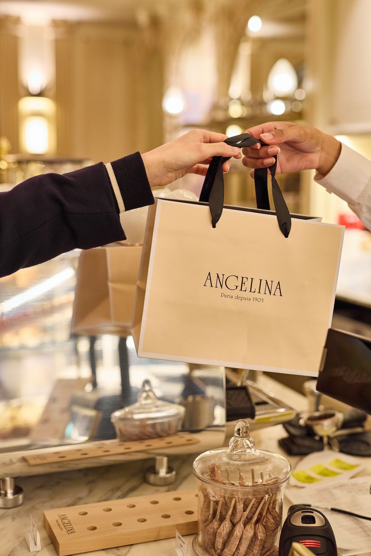 Hand passing a beige shopping bag with Angelina Paris logo inside a tea room or pastry shop environment.