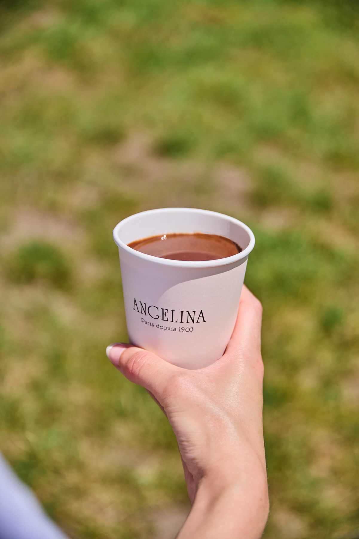 Hand holding a white cup of hot chocolate with the Angelina logo, outdoors on a grass background.