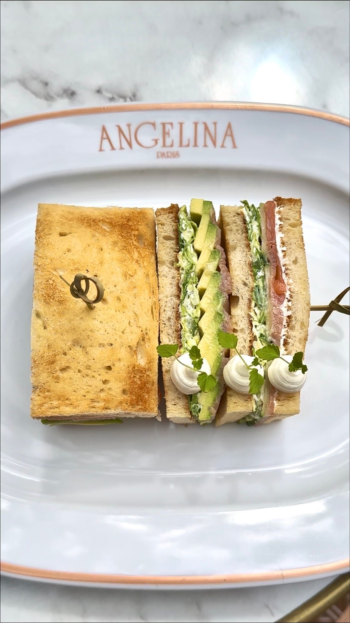 Club sandwich served on a white plate with Angelina Paris logo, featuring layers of bread, avocado, cream, and meat.