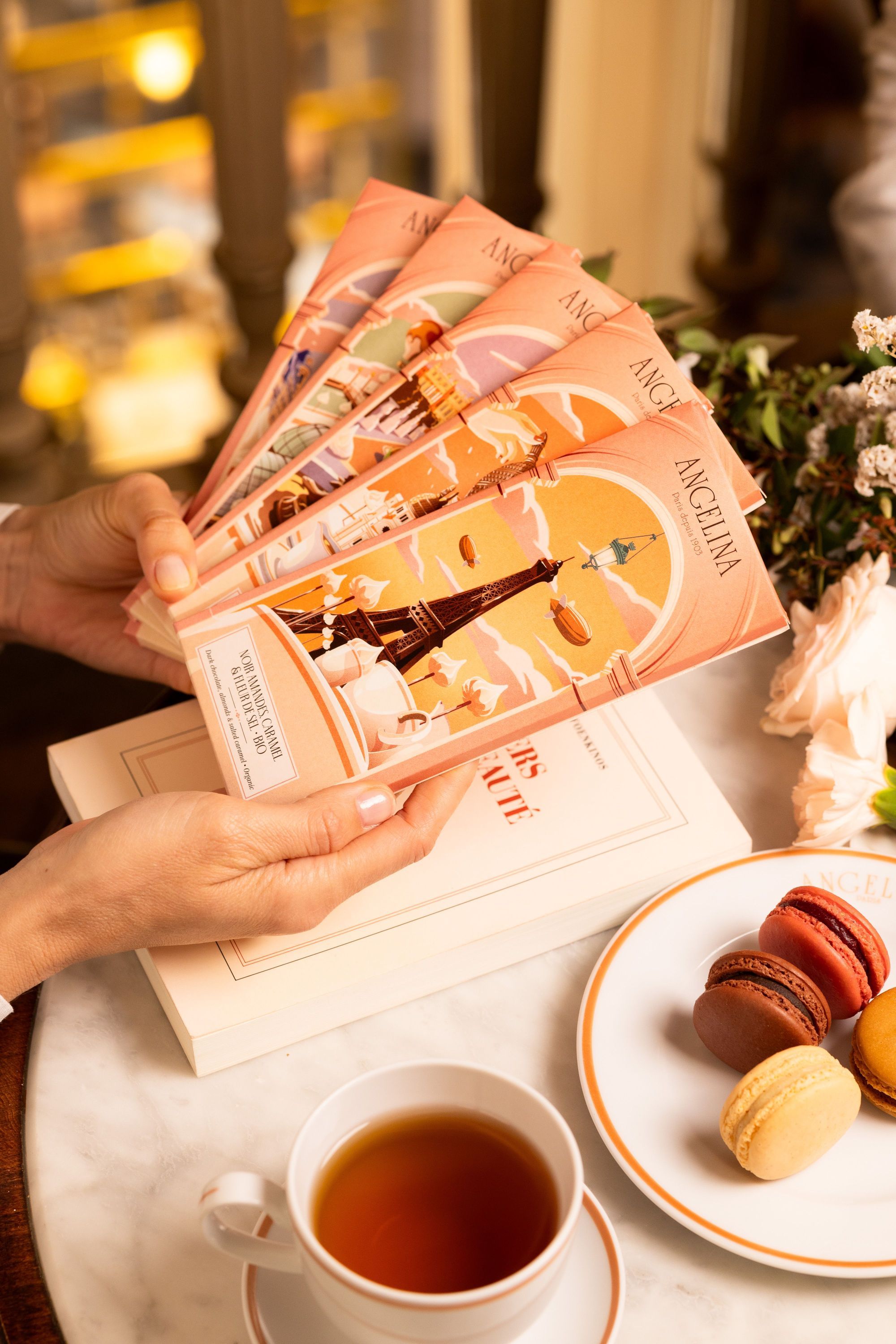 Several pastel-colored Boutique Angelina Paris menus with illustrations, held over a marble table with tea and macarons.