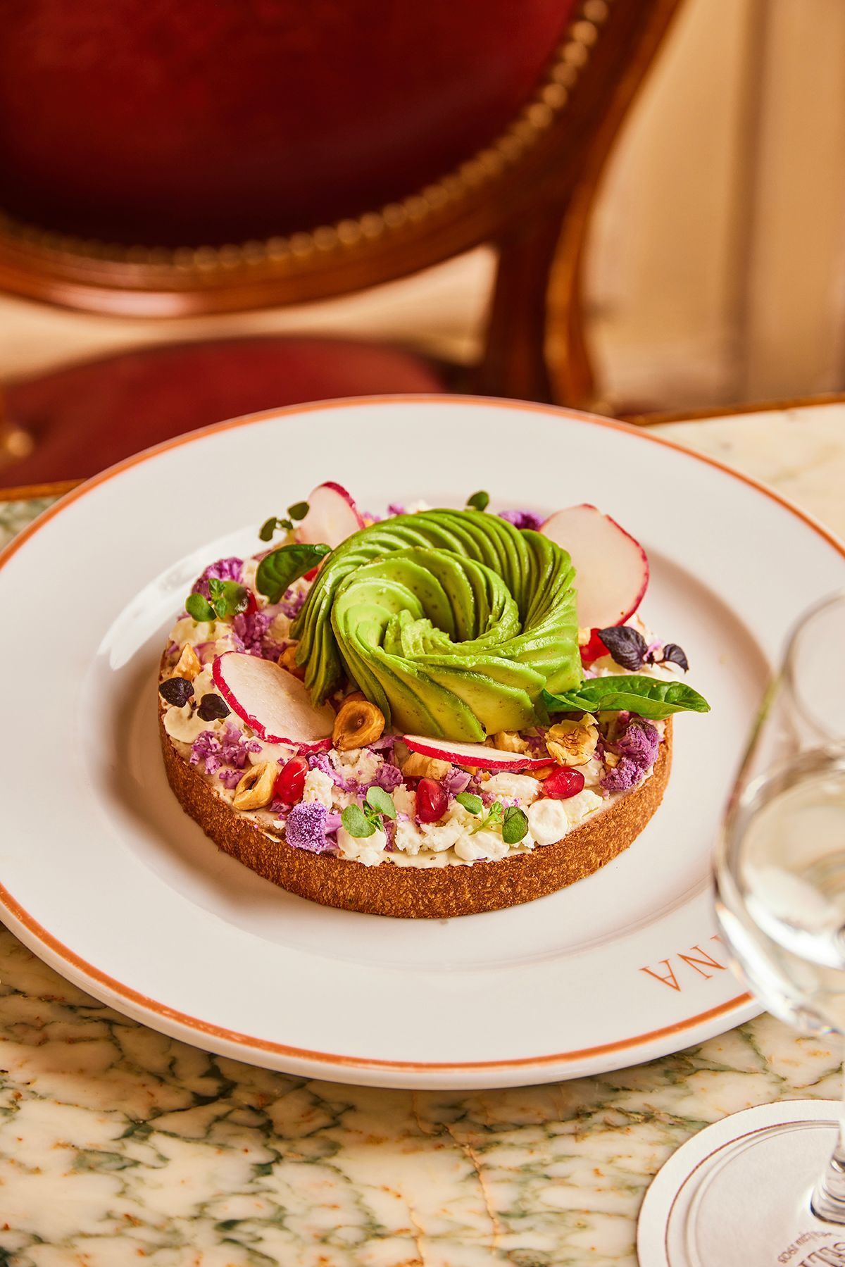 Tarte fine décorée d'avocat tranché, noix, fleurs comestibles et graines, servie sur une assiette de pâtisserie.