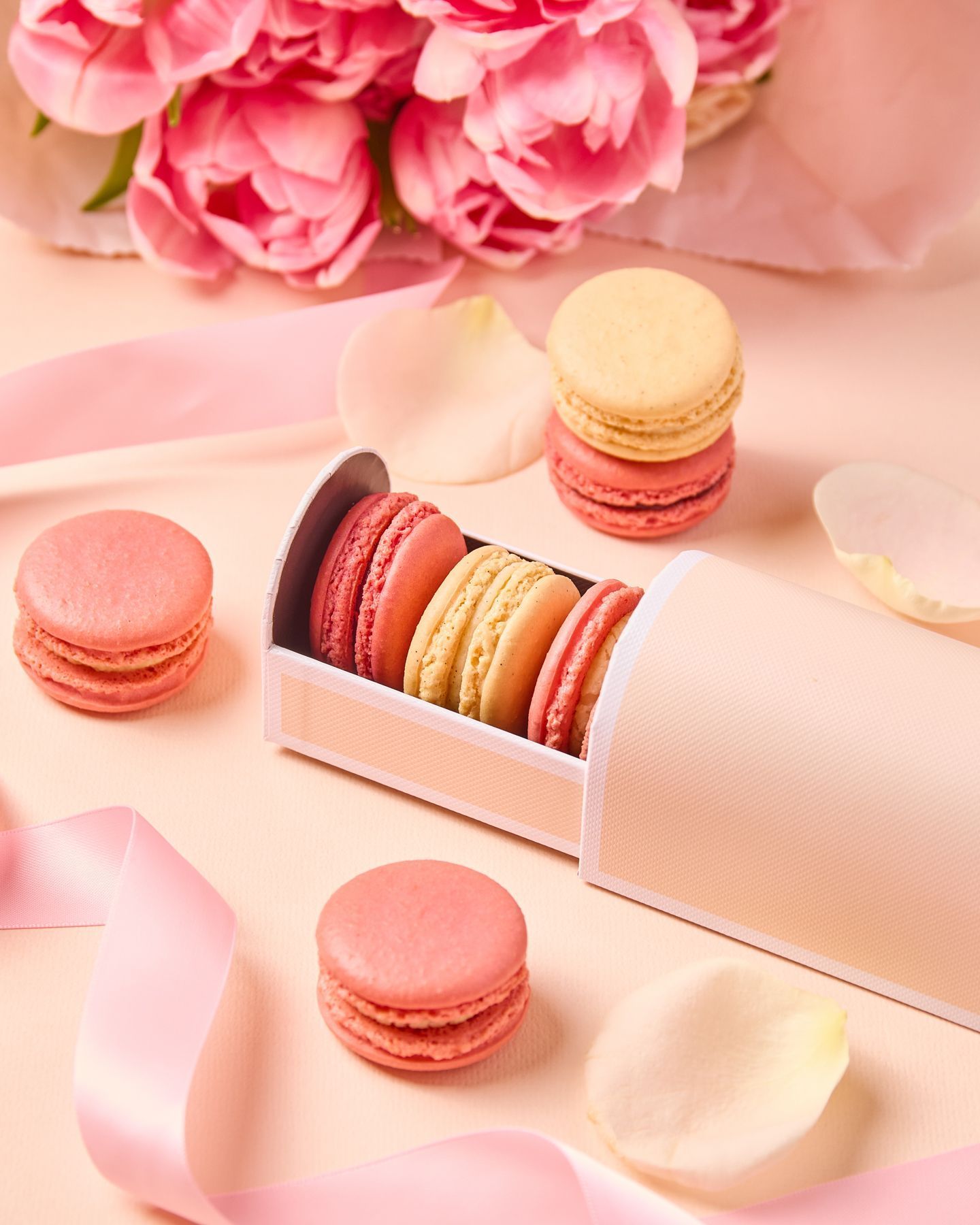 Colorful macarons in a box surrounded by pink roses, petals, and ribbons on a soft pink background.