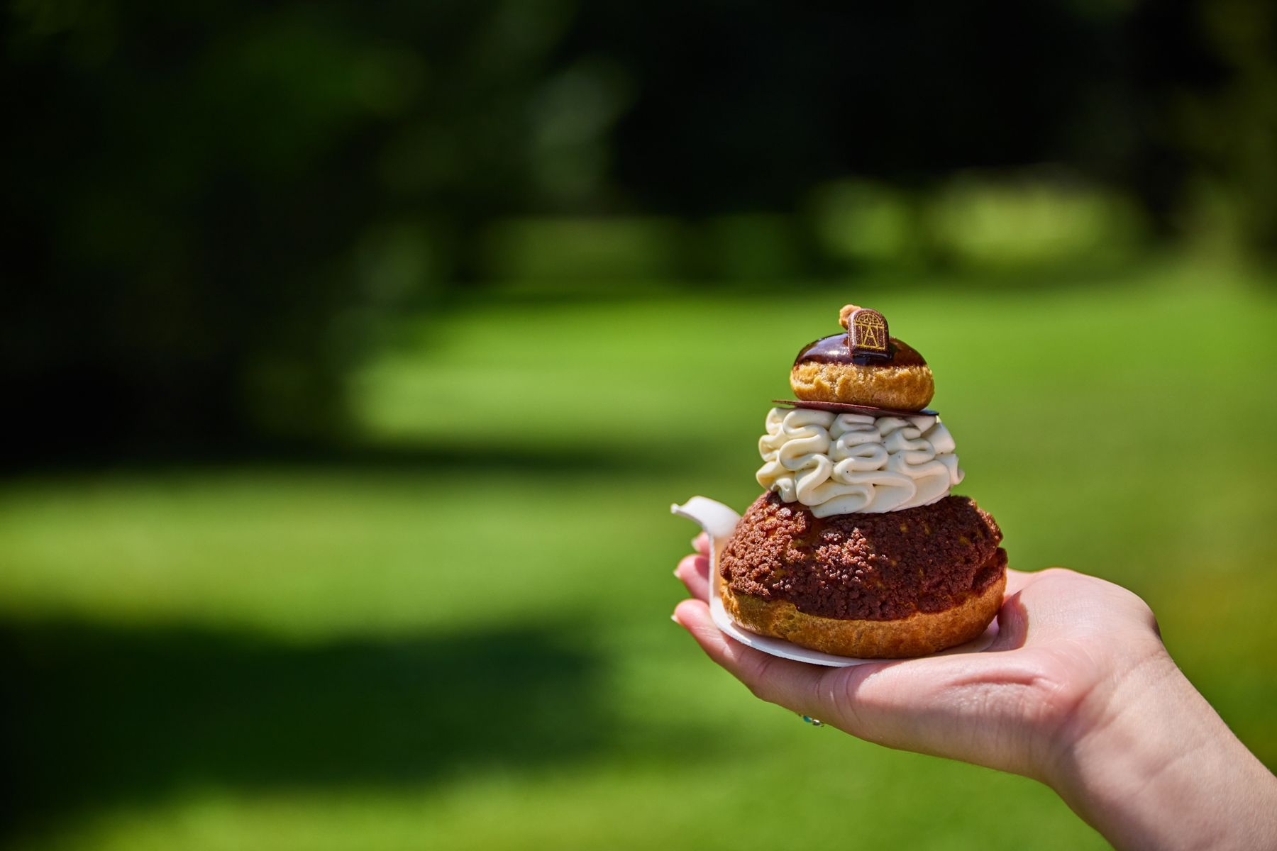 Delicate Mont-Blanc pastry topped with chocolate piece, held in hand against a blurred lush green background outdoors.