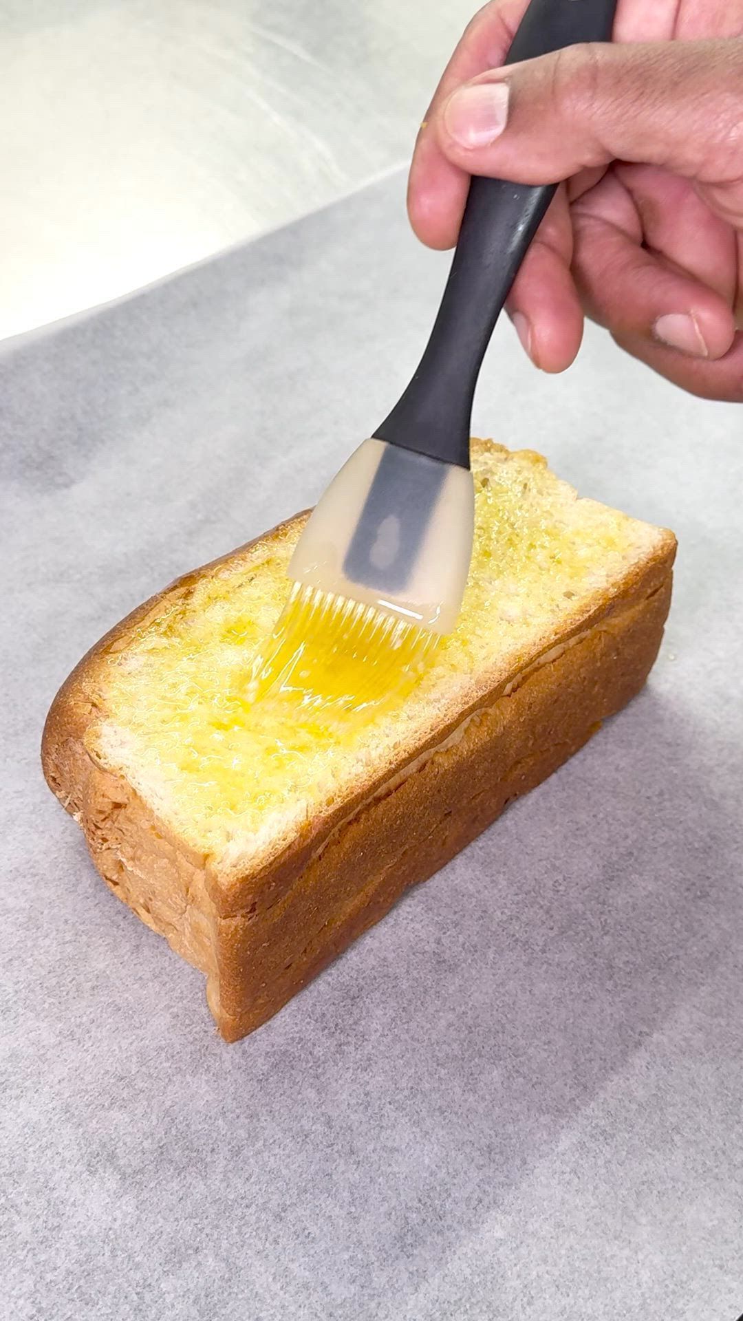 Un toast beurré sur une feuille de papier cuisson avec un pinceau en silicone appliquant du beurre fondu.