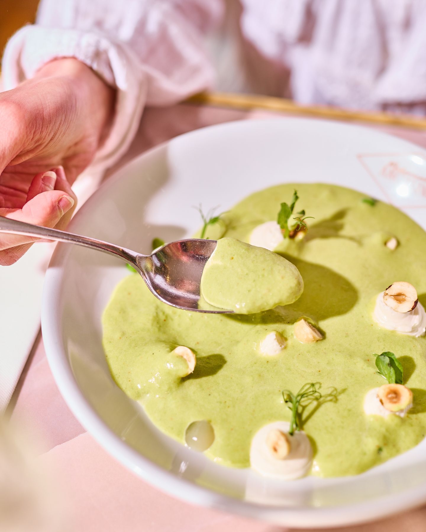 Velouté vert servi dans une assiette blanche avec des noisettes et des herbes, accompagné d'une cuillère en main