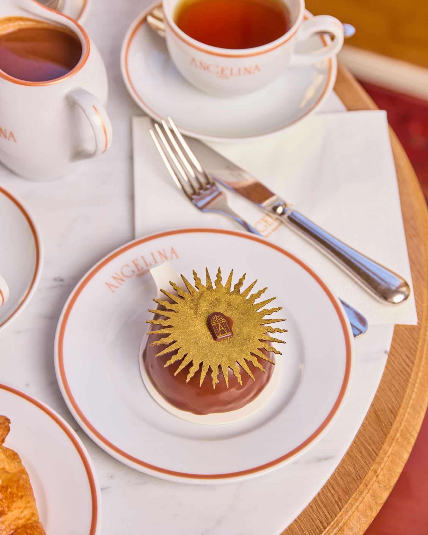 Pastry with golden decoration served on a white Angelina Paris plate with a cup of tea and utensils on a wooden table.