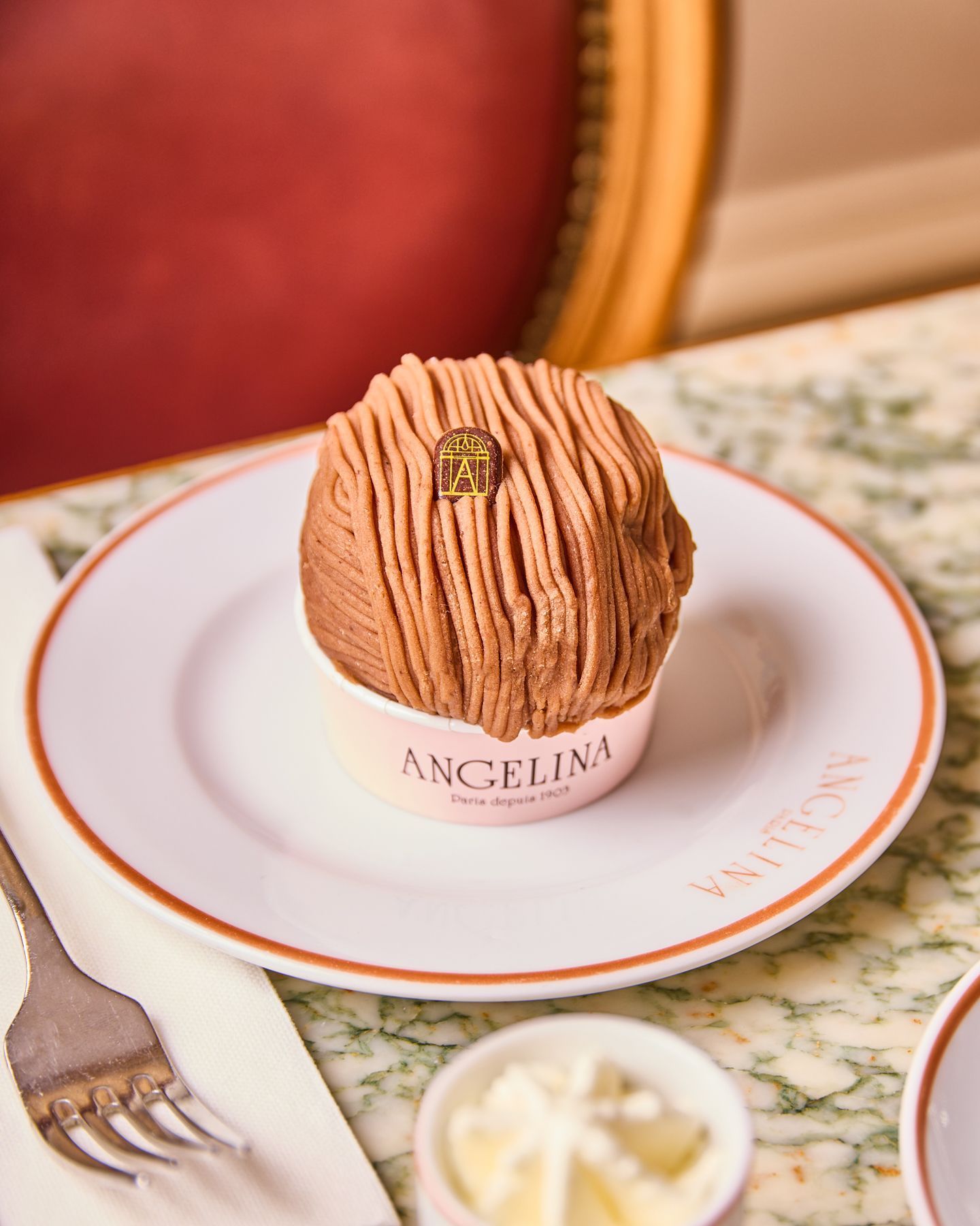 Mont-Blanc dessert served on a white plate with Angelina branding, placed on a marble table with a red background.