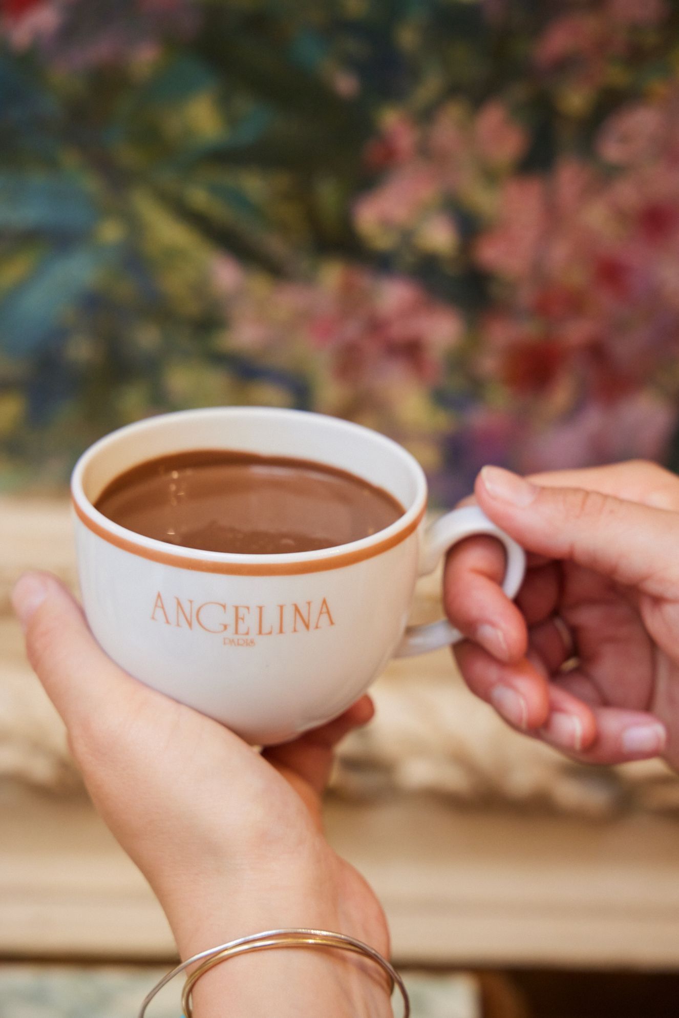 Hands holding a cup of hot chocolate with Angelina Paris logo, set against a blurred floral background.